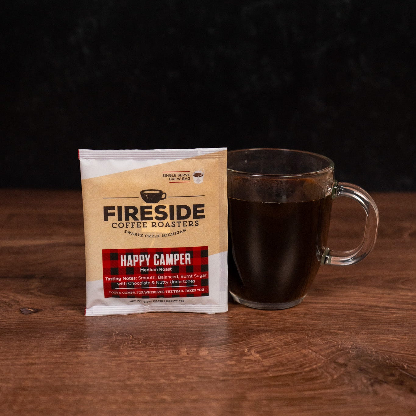 Fireside Coffee Masters Happy Camper coffee package with a glass mug of coffee on a wooden surface.